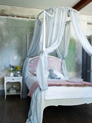 Children’s white four-poster bed with blue canopy, pink headboard, and soft pastel bedding in a rustic room.